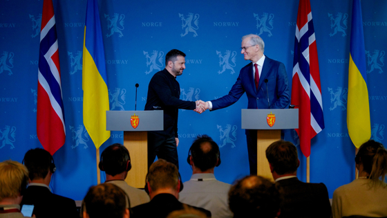 Støre og Zelenskyj tar hverandre i hendene på podiet hvor pressekonferansen holdes. Norske og ukrainske flagg. Blå bakgrunn med riksløver og "Norway".